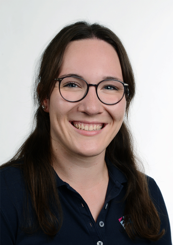 Porträt einer lächelnden Frau mit langen, braunen Haaren, die ein dunkelblaues Hemd und eine Brille trägt. Sie steht vor einem neutralen Hintergrund.