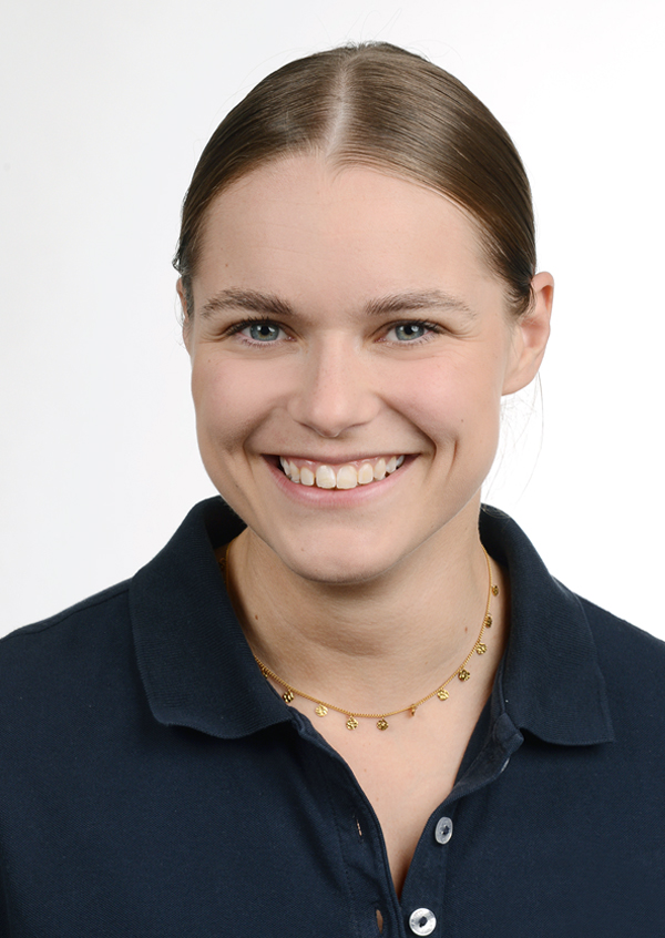 Porträt einer lächelnden Frau mit zugebundenem Haar, die ein blaues Shirt trägt. Der Hintergrund ist neutral und hell.
