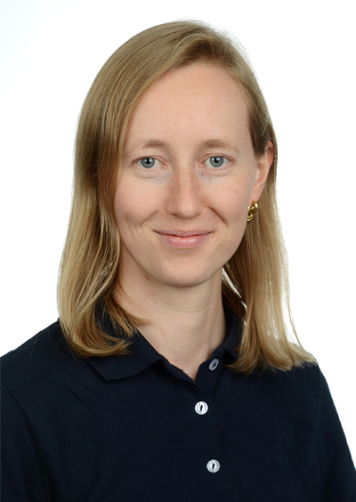 Porträt einer lächelnden Frau mit mittellangen, blonden Haaren, die ein dunkelblaues Hemd trägt. Sie steht vor einem neutralen Hintergrund.