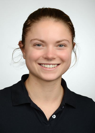 Merle Woyczechowski Porträt einer lächelnden Frau mit zugebundenem Haar, die ein blaues Shirt trägt. Der Hintergrund ist neutral und hell.