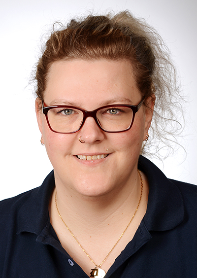 Porträt einer lächelnden Frau mit zugebundenem Haar, die ein blaues Shirt trägt. Die Frau trägt eine Brille. Der Hintergrund ist neutral und hell.