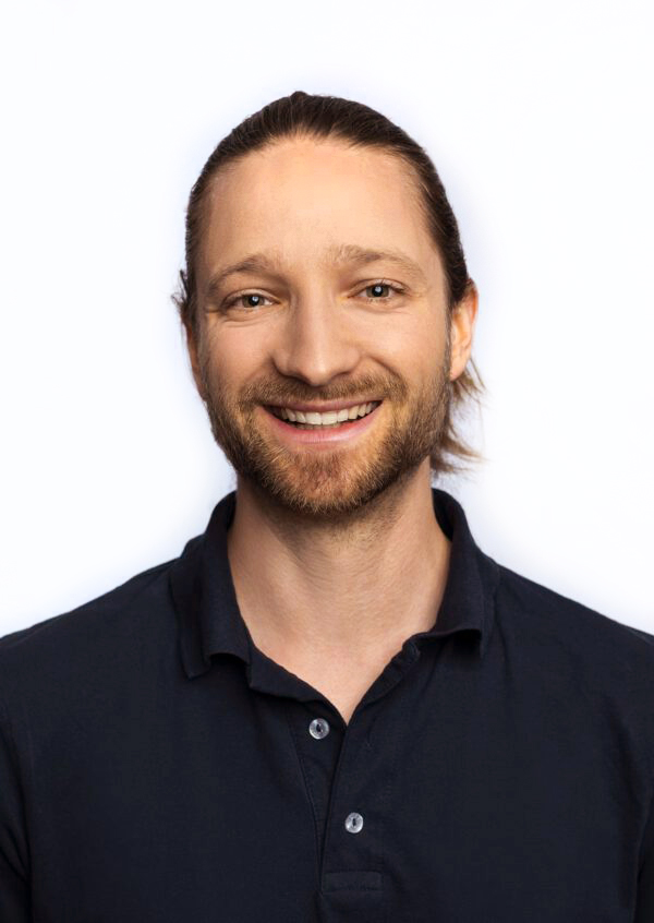 Sten Radü Lächelnder Mann mit zurückgebundenen Haaren in einem schwarzen Poloshirt vor einem hellen Hintergrund.