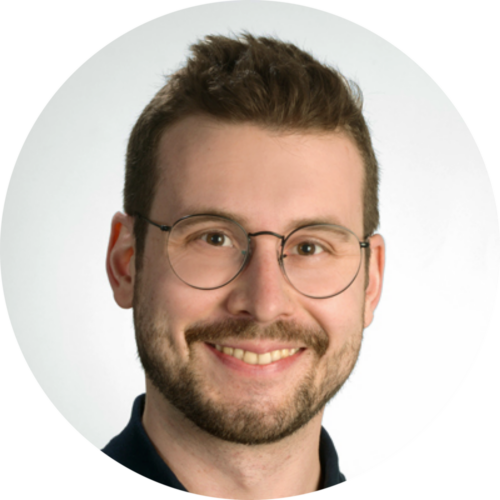 Yannick Wilts Rundes Porträtfoto eines lächelnden Mannes mit Bart und runder Brille vor einem hellen Hintergrund.